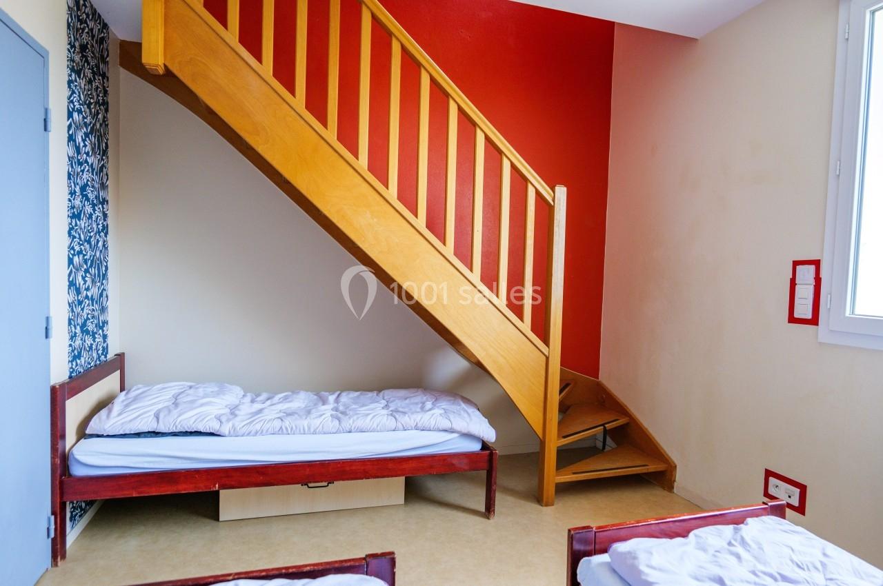 Chambre simple avec lits individuels, escalier en bois, mur rouge et fenêtre apportant de la lumière naturelle.