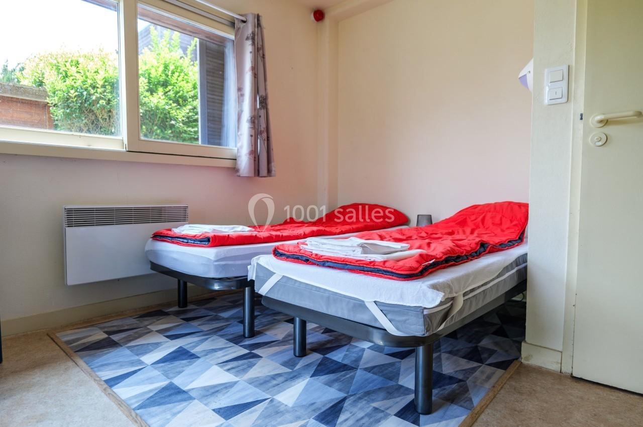 Deux lits simples avec draps rouges dans une chambre lumineuse, dotée d'une fenêtre et d'un tapis géométrique.