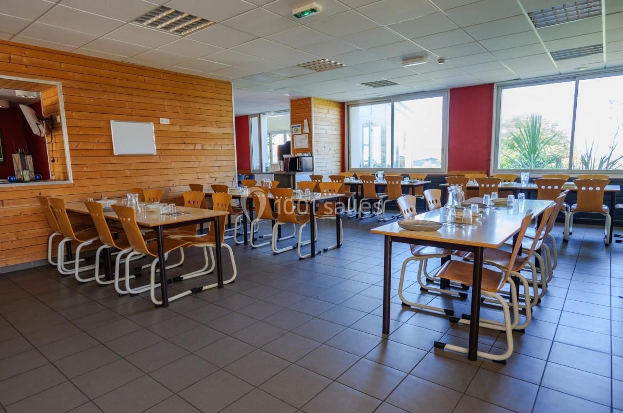 Salle de restauration lumineuse avec tables en bois, chaises alignées et grandes fenêtres donnant sur l'extérieur.