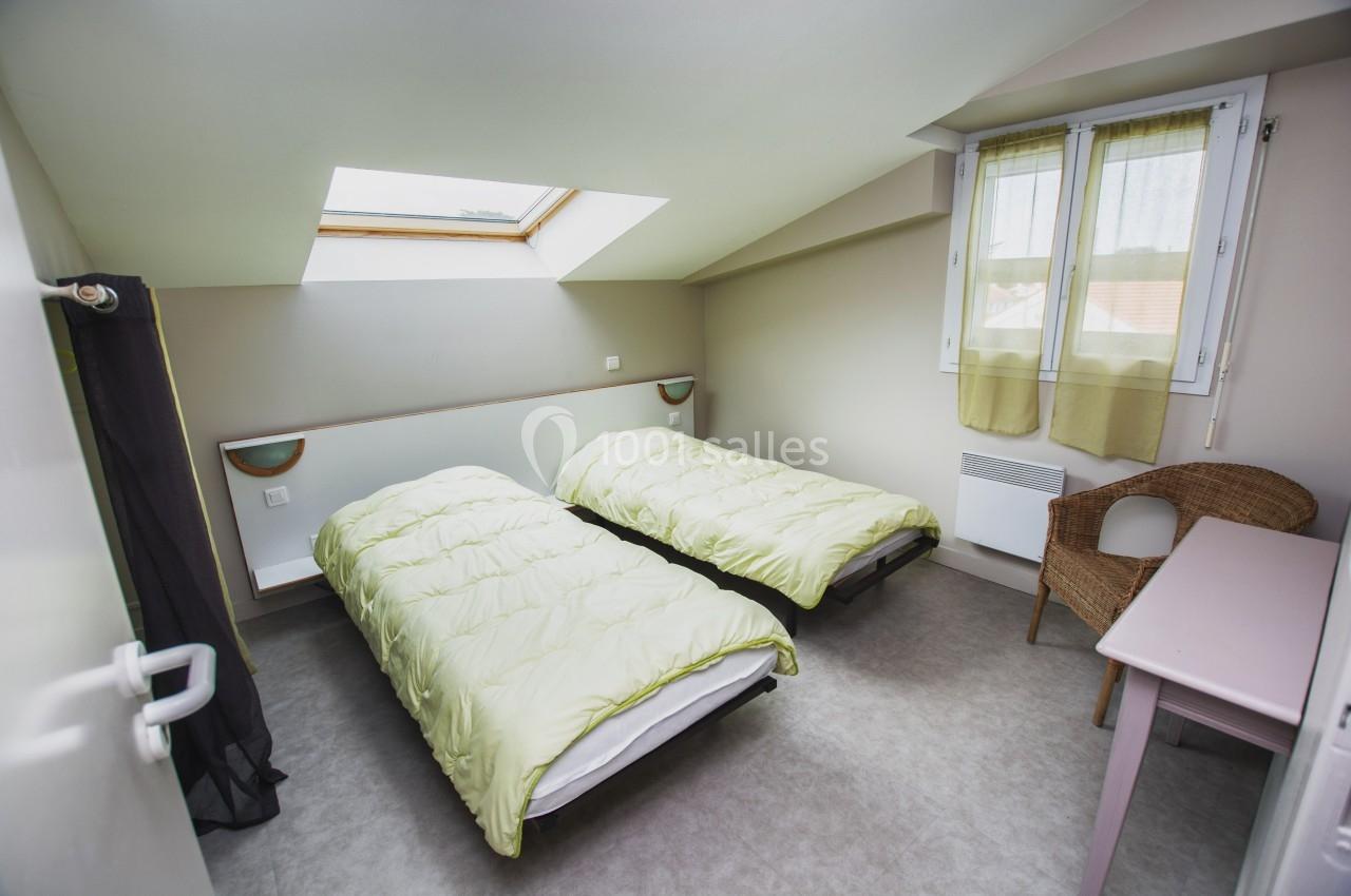 Chambre mansardée avec deux lits simples, fenêtre de toit, bureau, chaise en osier et lumière naturelle.