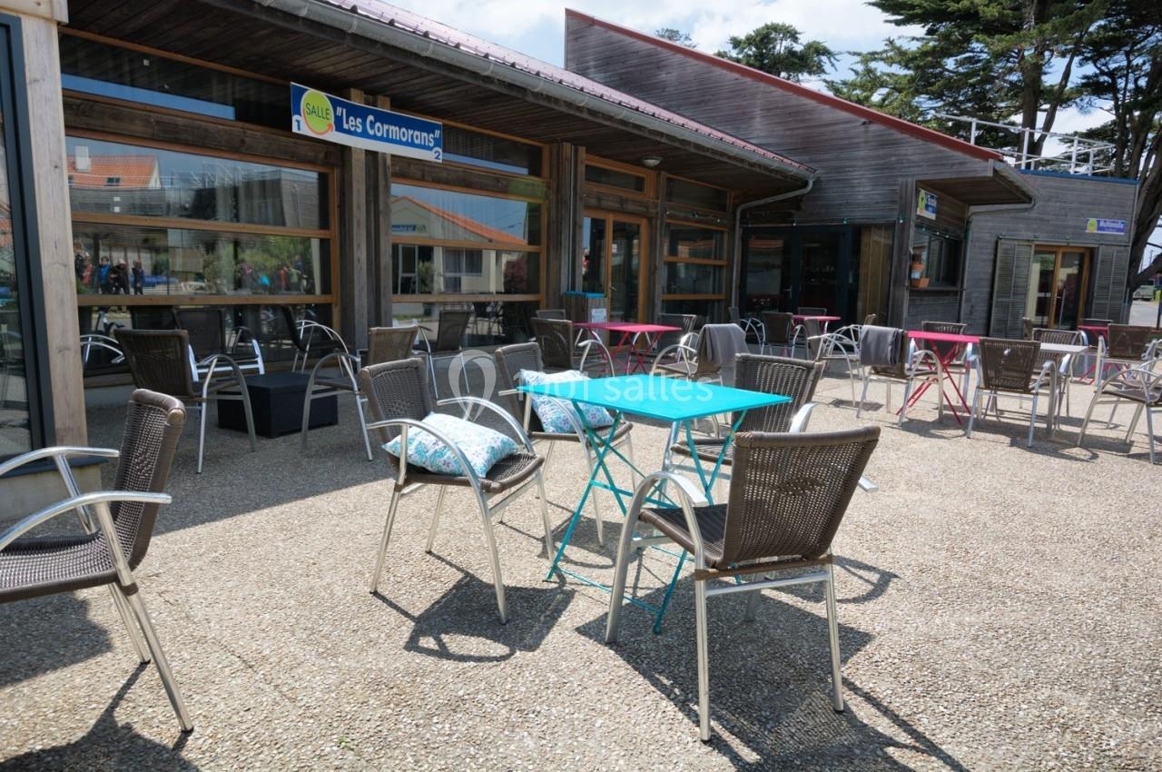 Terrasse extérieure avec tables et chaises en métal, devant un bâtiment en bois avec de grandes fenêtres.