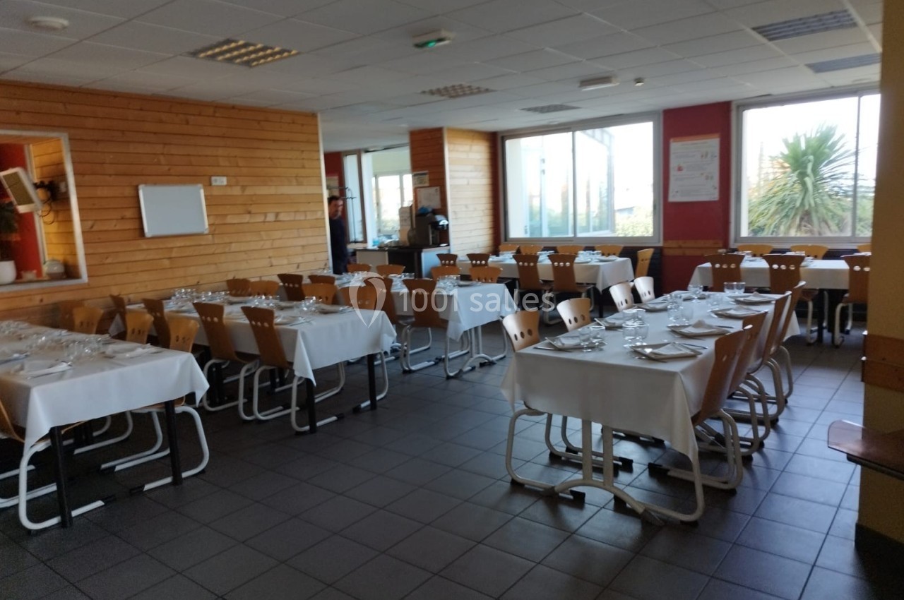 Salle de restaurant avec des tables dressées, nappes blanches et chaises en bois, éclairée par des fenêtres.