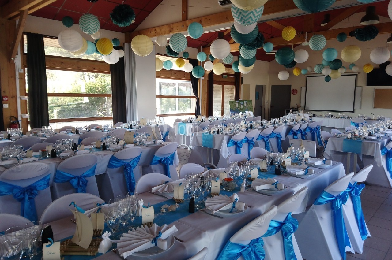 Salle décorée pour un événement avec des tables dressées, des chaises ornées de nœuds bleus et des lanternes suspendues.