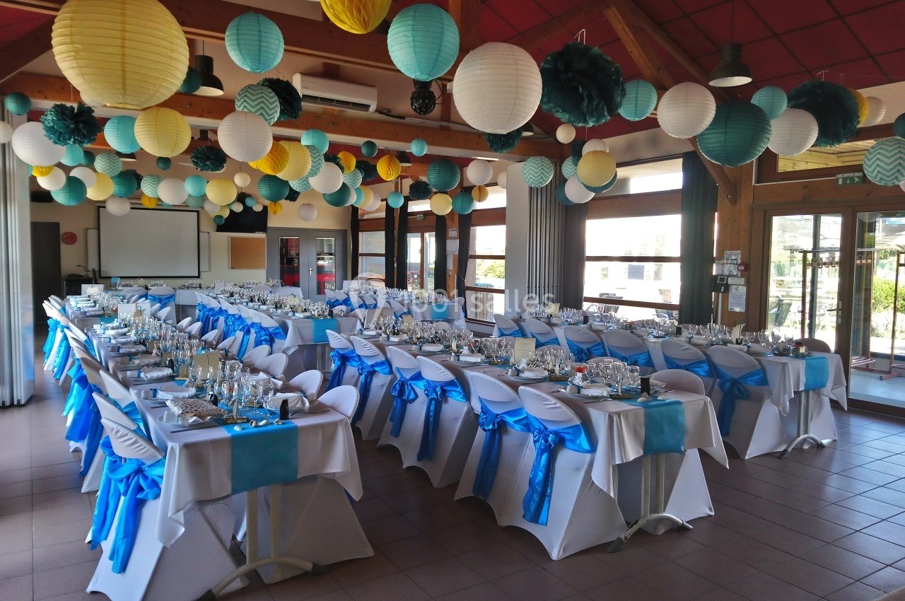 Salle décorée pour un événement, avec des tables dressées et des chaises ornées de nœuds bleus, sous des lanternes…