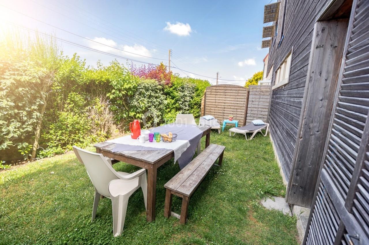 Table en bois avec bancs et chaises sur une pelouse, entourée de haies, près d'une maison en bois sous un ciel ensoleillé.