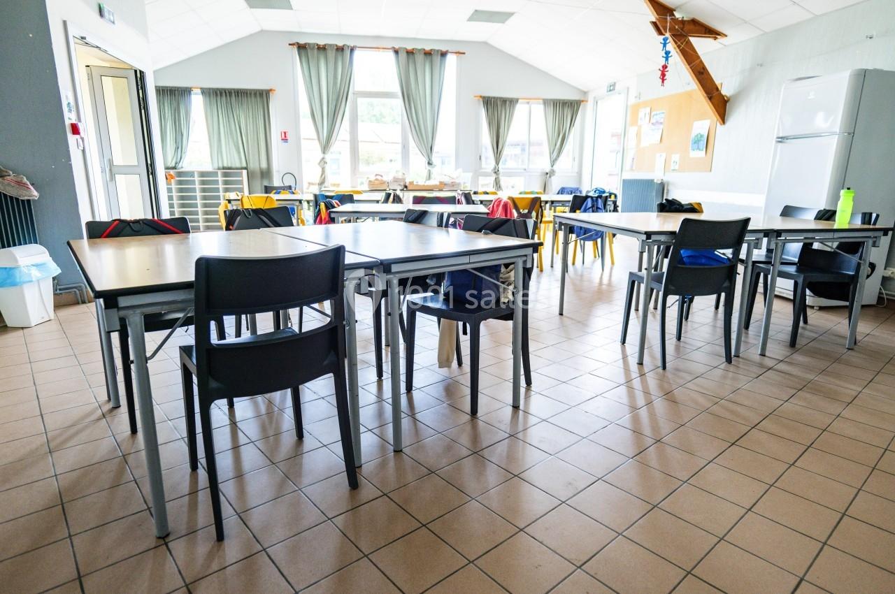 Salle lumineuse avec des tables et des chaises noires, rideaux ouverts sur des fenêtres, et un réfrigérateur au fond.