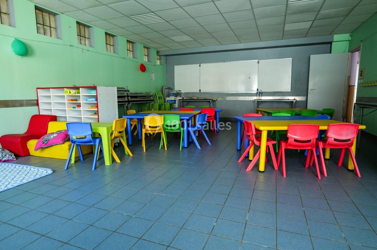 Salle de classe colorée avec des tables et chaises pour enfants, étagères de rangement et tableau blanc au fond.