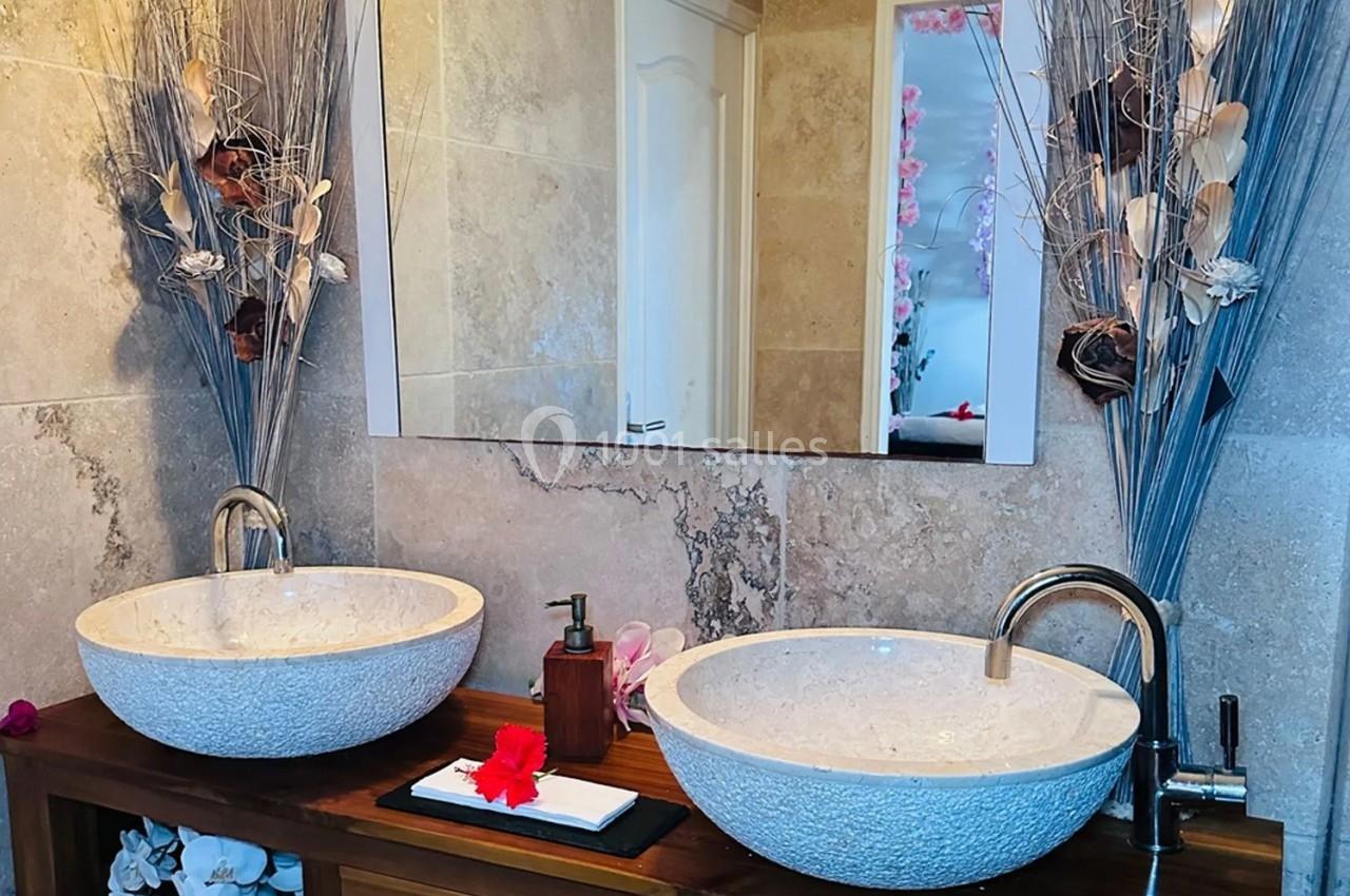 Deux vasques en pierre blanche sur un meuble en bois, décorées avec des fleurs et des branches sèches, devant un miroir.