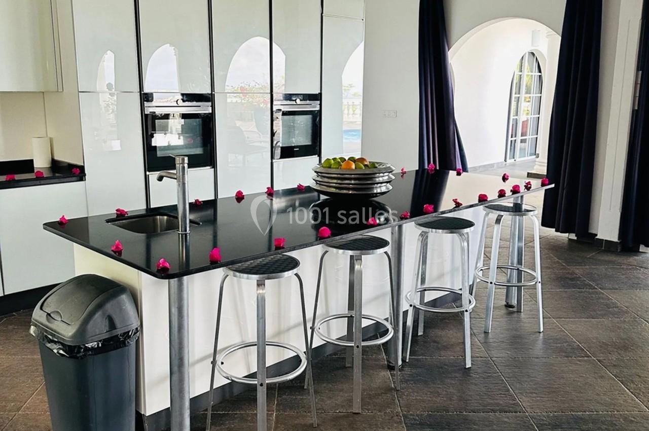 Cuisine moderne avec îlot central noir, tabourets transparents, évier intégré et décorations de pétales roses.