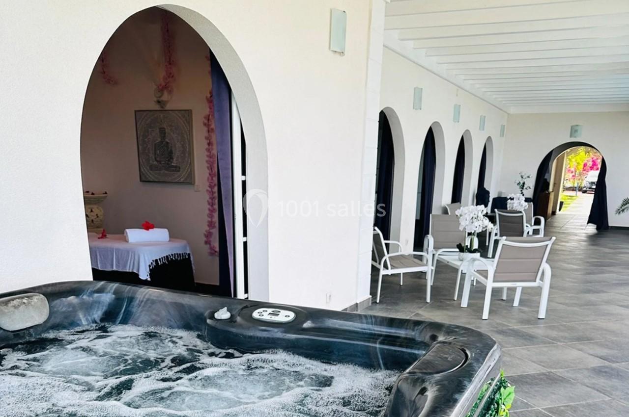 Vue d'une terrasse couverte avec jacuzzi, chaises blanches, table et décoration florale dans un cadre lumineux.