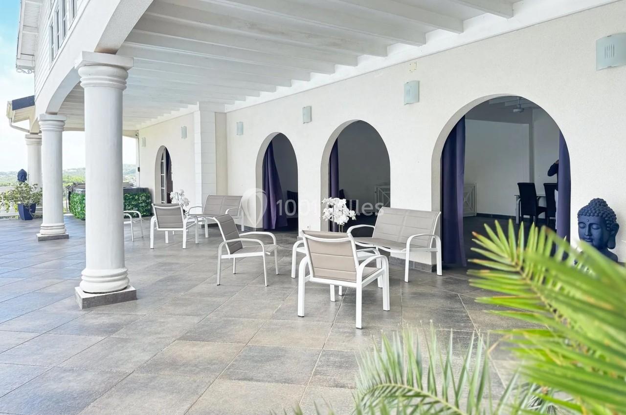 Terrasse couverte avec mobilier de jardin blanc, arches ouvertes et décorations, entourée de plantes.