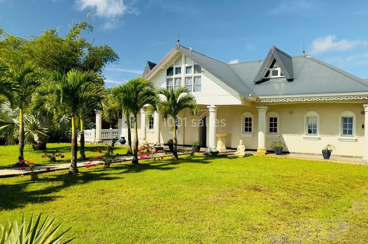 Maison blanche avec toit gris entourée de palmiers et d'un jardin verdoyant sous un ciel bleu clair.