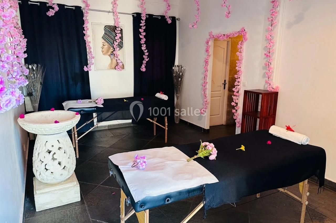 Salle de massage avec deux tables recouvertes de draps noirs, décorée de guirlandes roses et d'une peinture murale.