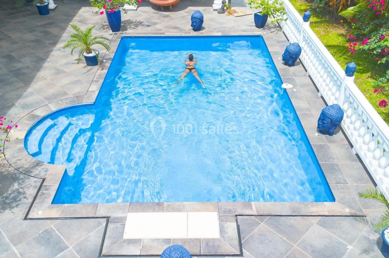 Vue aérienne d'une piscine rectangulaire avec une personne nageant, entourée de plantes et de décorations bleues.