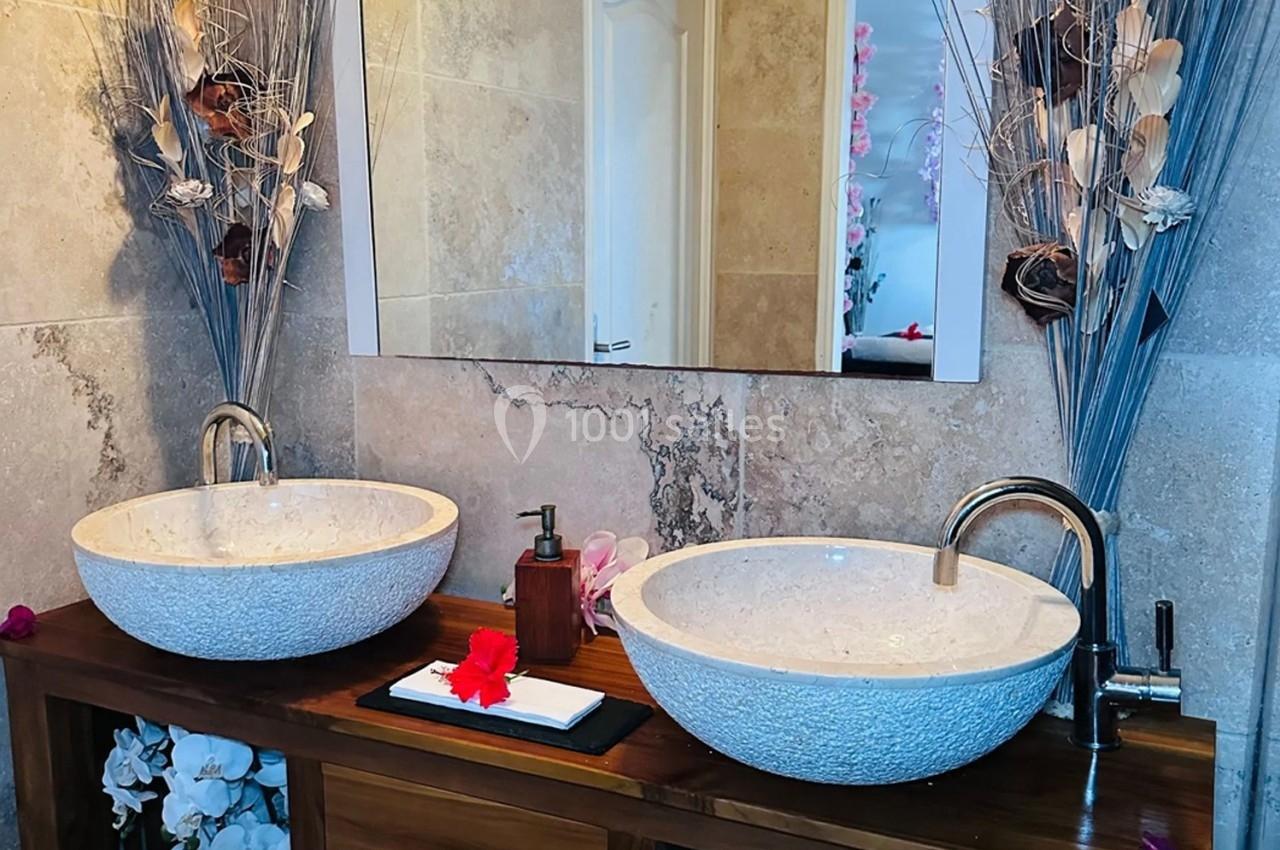 Double vasque en pierre sur meuble en bois, décorée de fleurs blanches et sèches, avec miroir et éclairage mural.