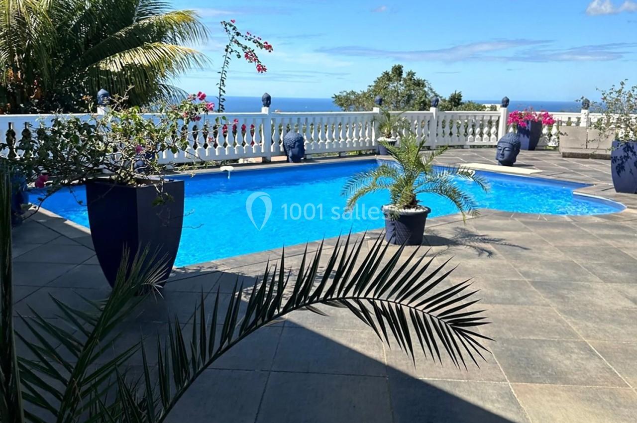Piscine extérieure entourée de plantes, avec vue sur la mer et un ciel bleu dégagé en arrière-plan.