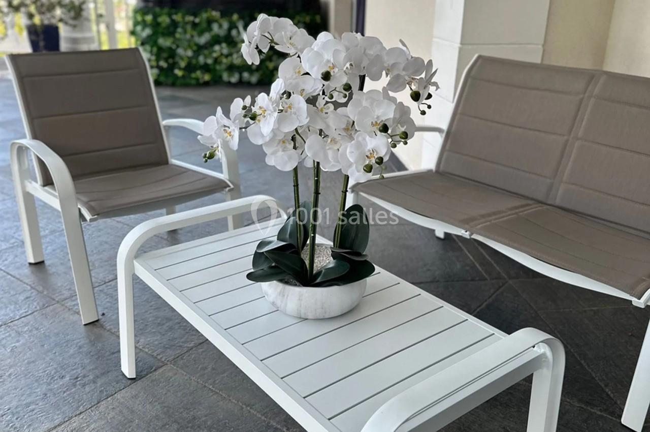 Table basse blanche avec une orchidée en pot, entourée de deux fauteuils beige sur une terrasse carrelée.