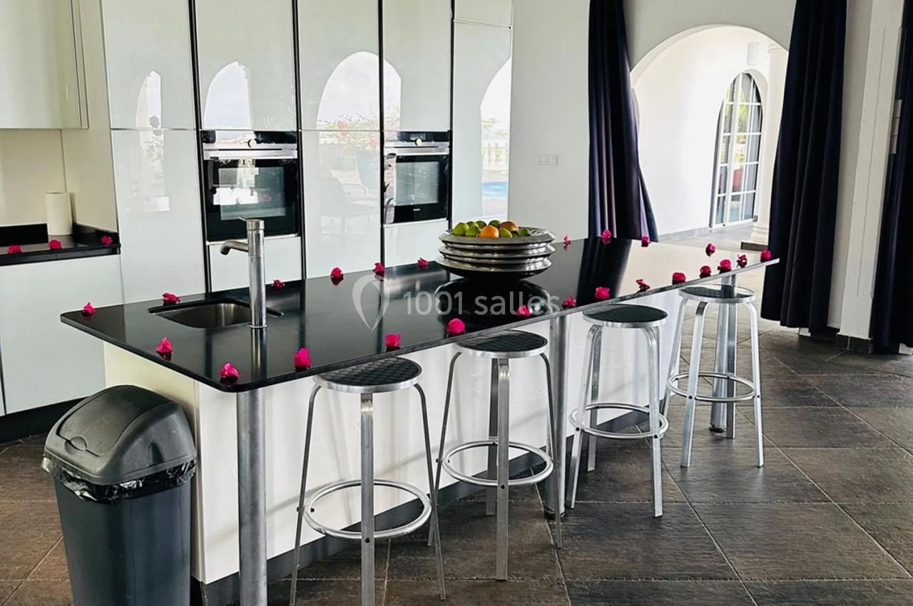 Cuisine moderne avec îlot central noir, tabourets métalliques, décorée de petites fleurs roses et éclairée par des fenêtres…