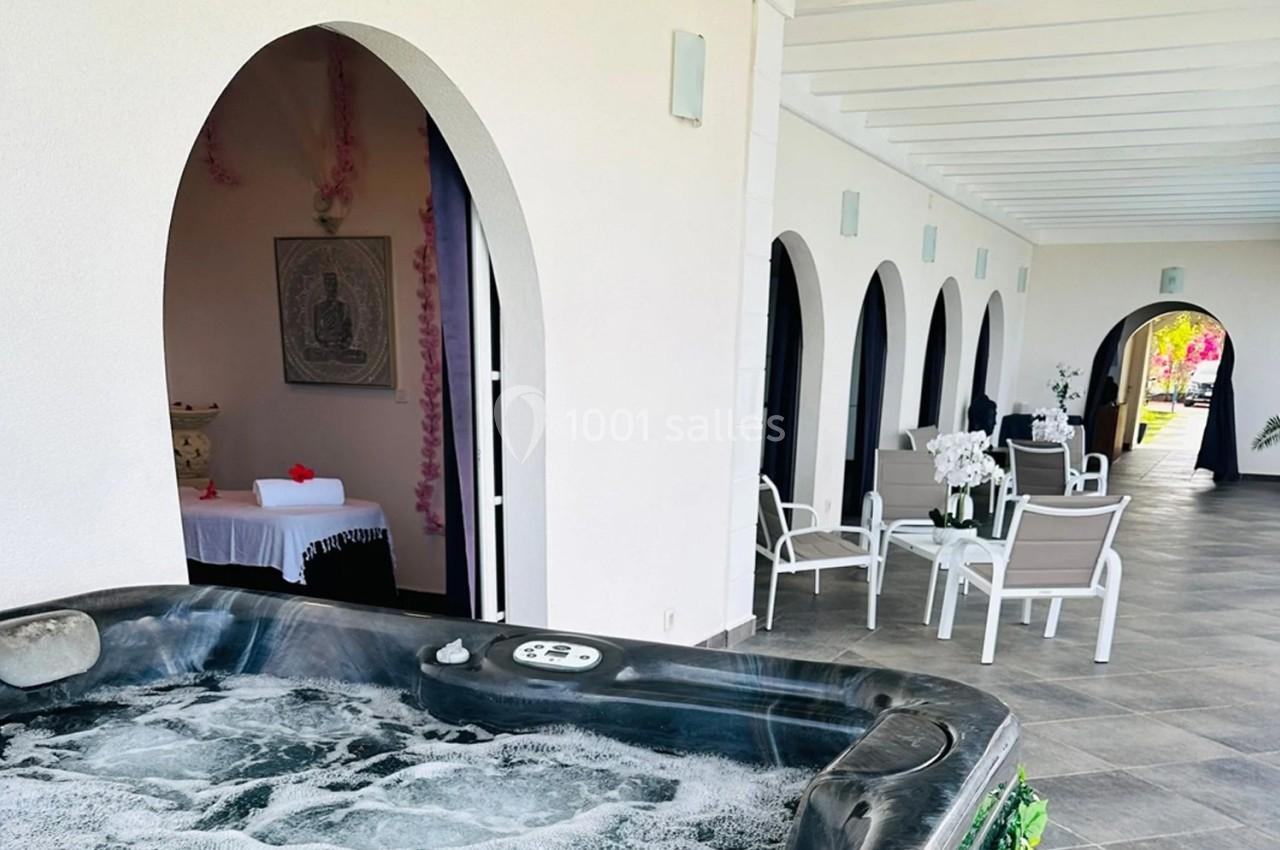 Bain à remous en premier plan avec terrasse couverte, chaises blanches et espace de massage en arrière-plan.