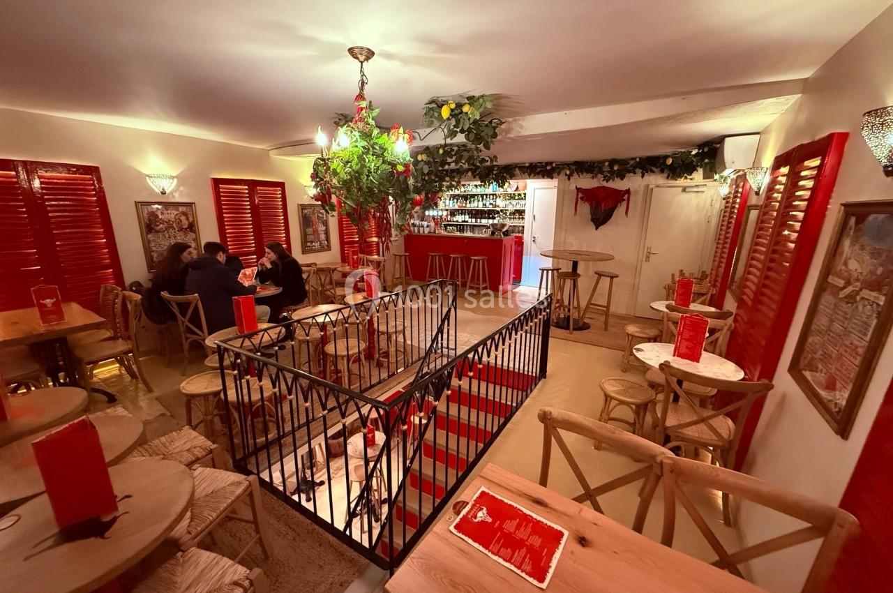 Salle de restaurant chaleureuse avec décoration rouge et végétale, tables en bois et bar en arrière-plan.
