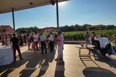 Un groupe de personnes échange autour d'un buffet en plein air, avec des serveurs et un paysage de vignoble en arrière-plan.