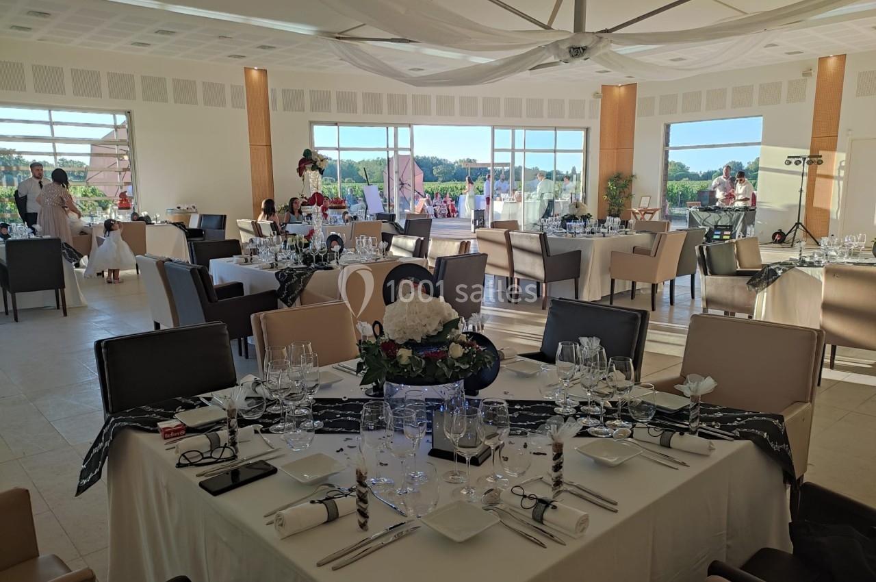 Salle de réception lumineuse avec tables décorées, nappes blanches et baies vitrées offrant une vue sur l'extérieur.