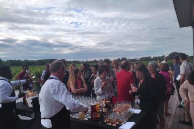 Un groupe de personnes échange autour d'un buffet en plein air, avec des serveurs et un paysage de vignoble en arrière-plan.