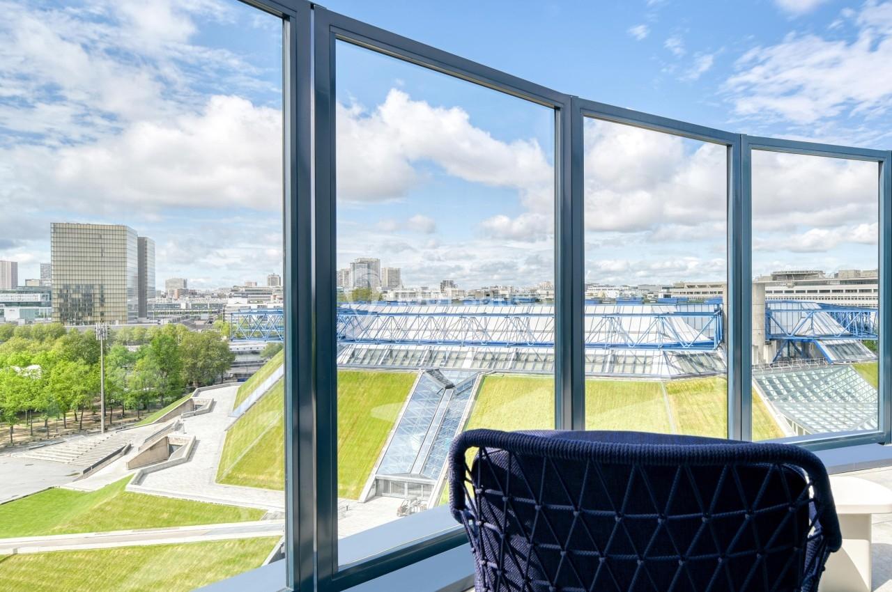 Vue depuis une fenêtre sur un toit végétalisé, des bâtiments modernes et un ciel partiellement nuageux.
