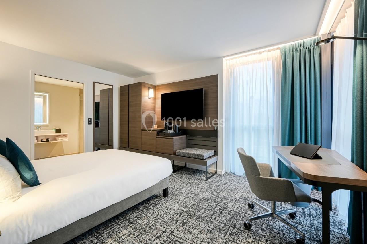 Chambre d'hôtel moderne avec lit double, bureau, télévision murale, salle de bain attenante et grandes fenêtres avec rideaux.