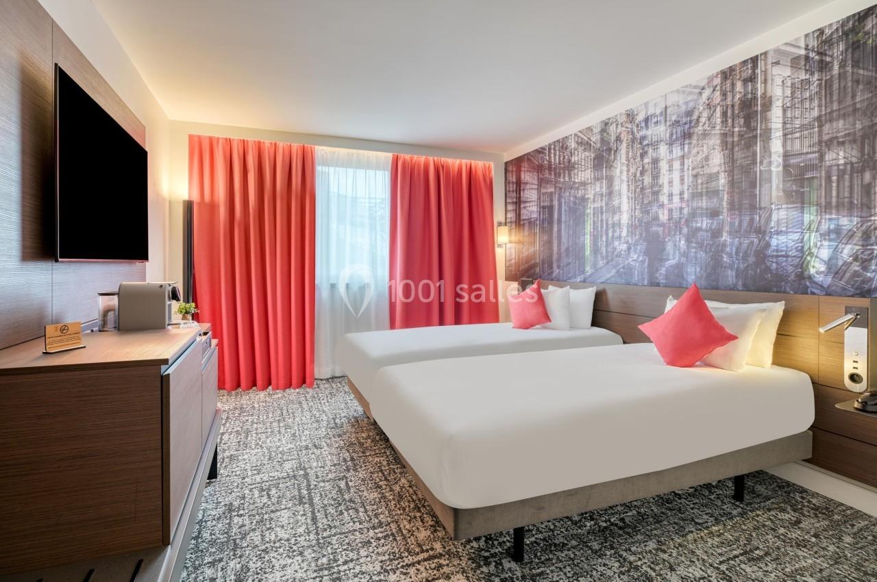 Chambre d'hôtel moderne avec deux lits simples, décoration murale urbaine, rideaux rouges et mobilier en bois clair.