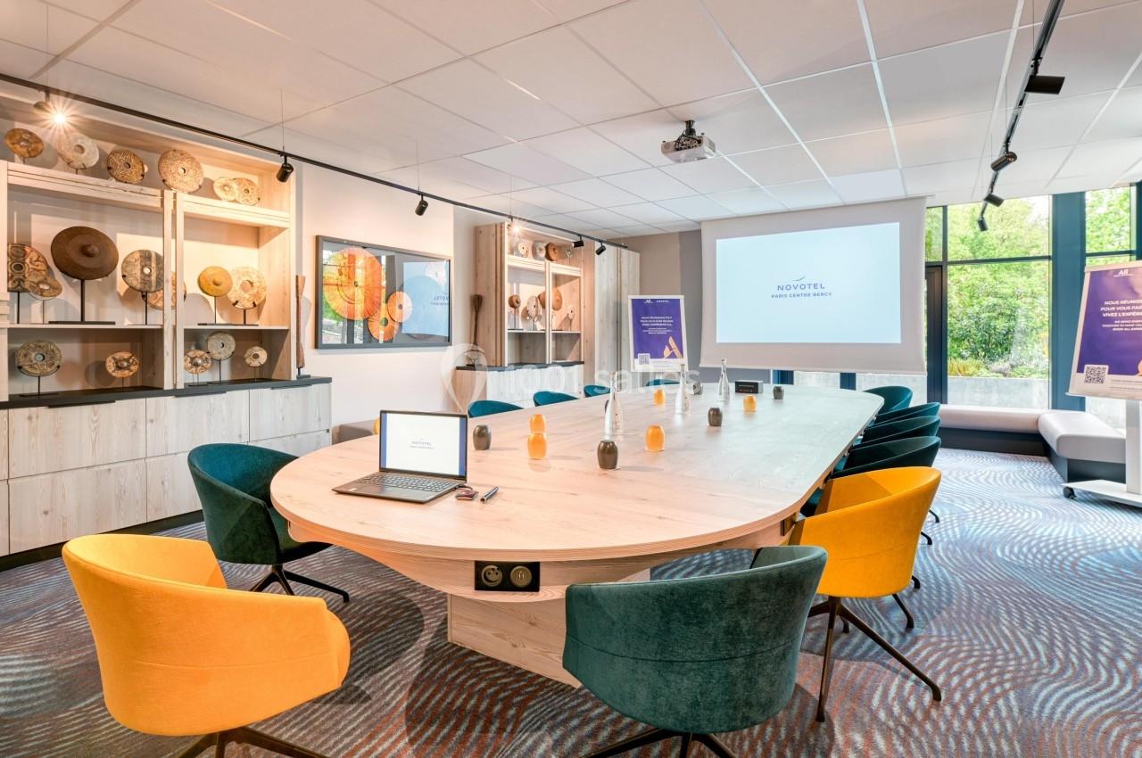 Salle de réunion lumineuse avec grande table ovale, chaises colorées, écran de projection et décorations murales.