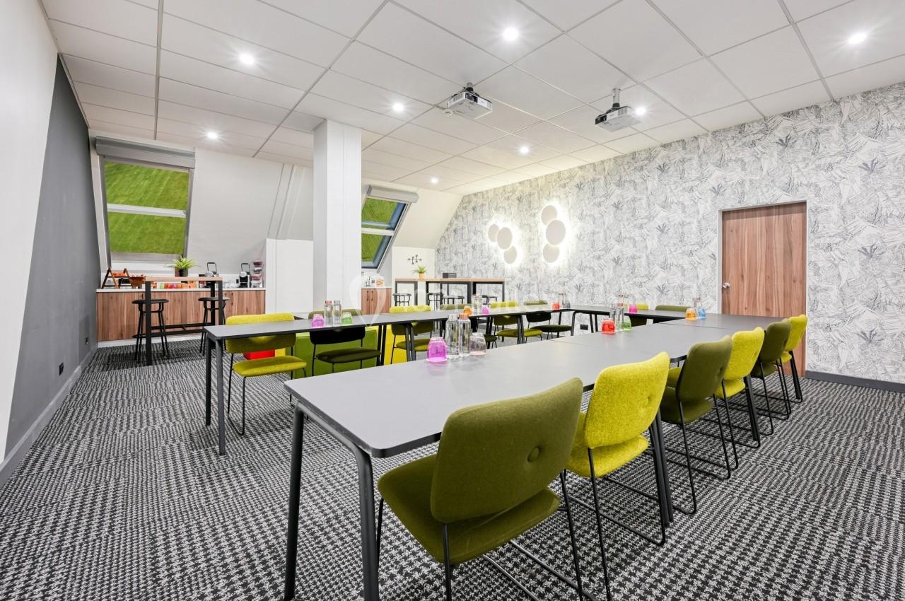 Salle de réunion lumineuse avec tables grises, chaises vertes, projecteurs au plafond et coin café en arrière-plan.