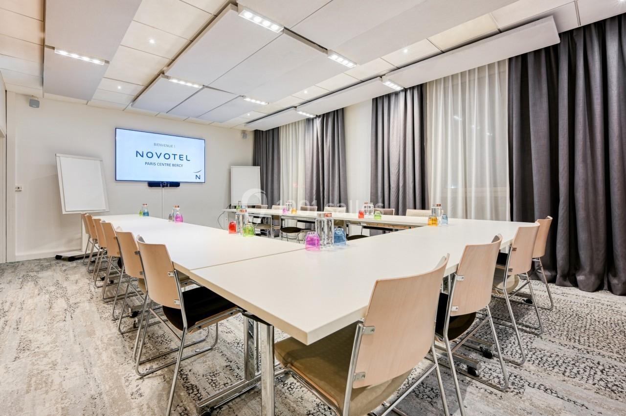 Salle de réunion lumineuse avec tables en configuration rectangulaire, chaises en bois clair et écran de présentation.