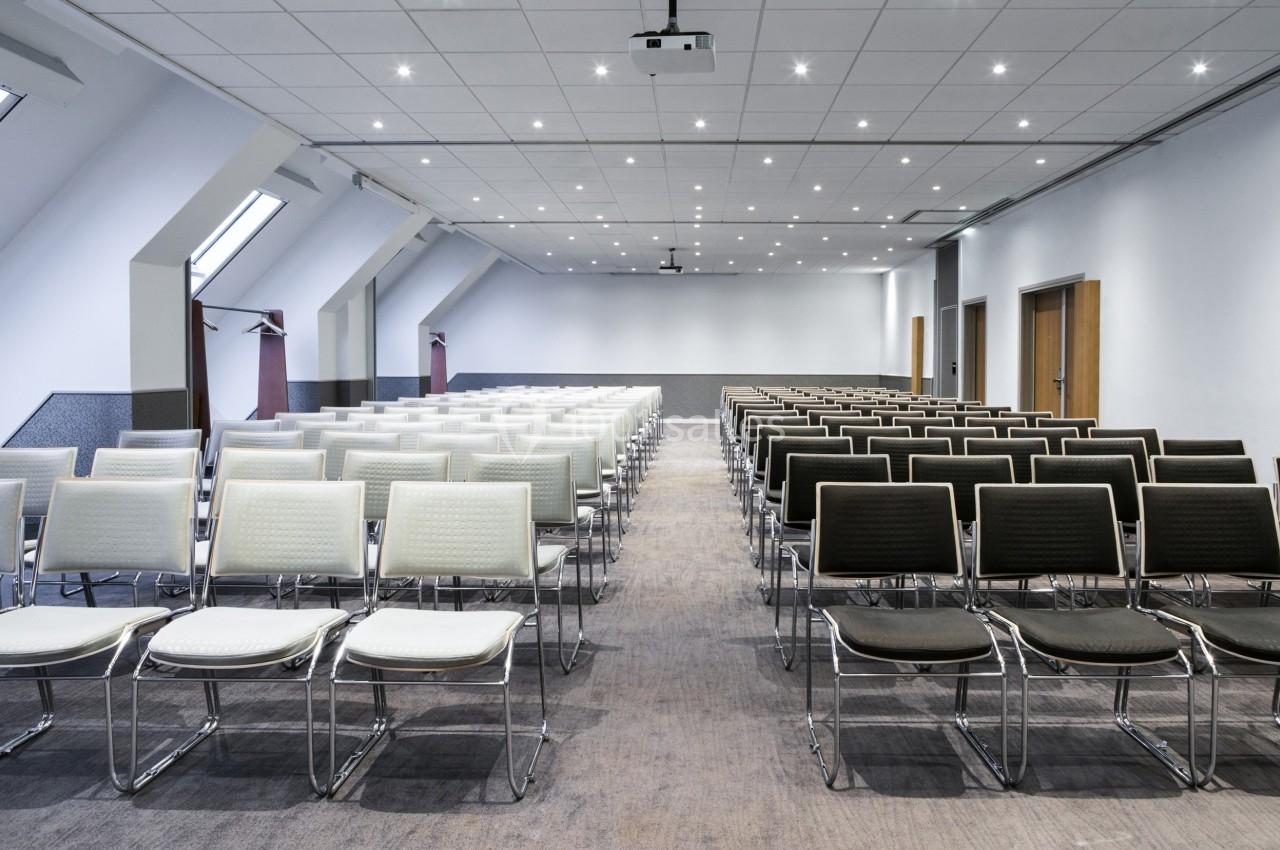 Salle de conférence vide avec des rangées de chaises blanches et noires, éclairée par des plafonniers et des fenêtres.