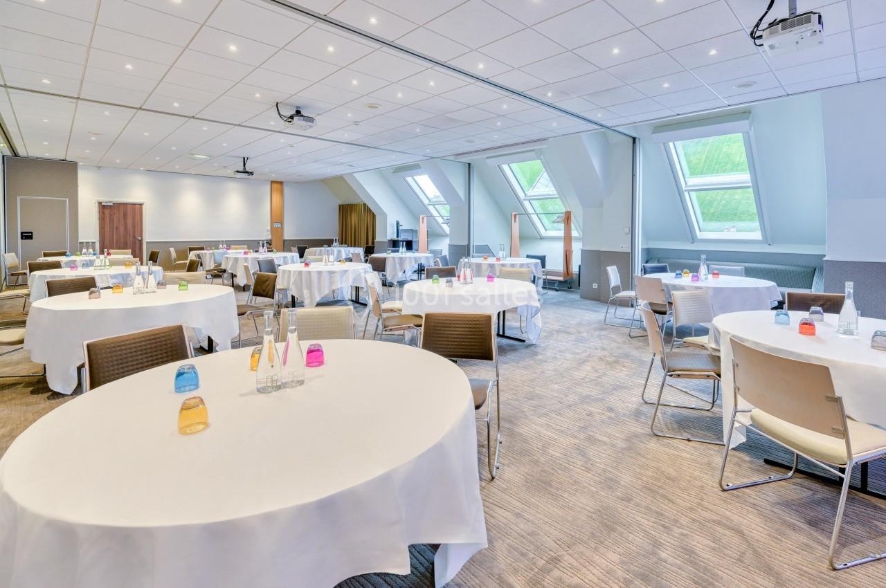 Salle de réunion lumineuse avec tables rondes dressées, chaises et grandes fenêtres laissant entrer la lumière naturelle.