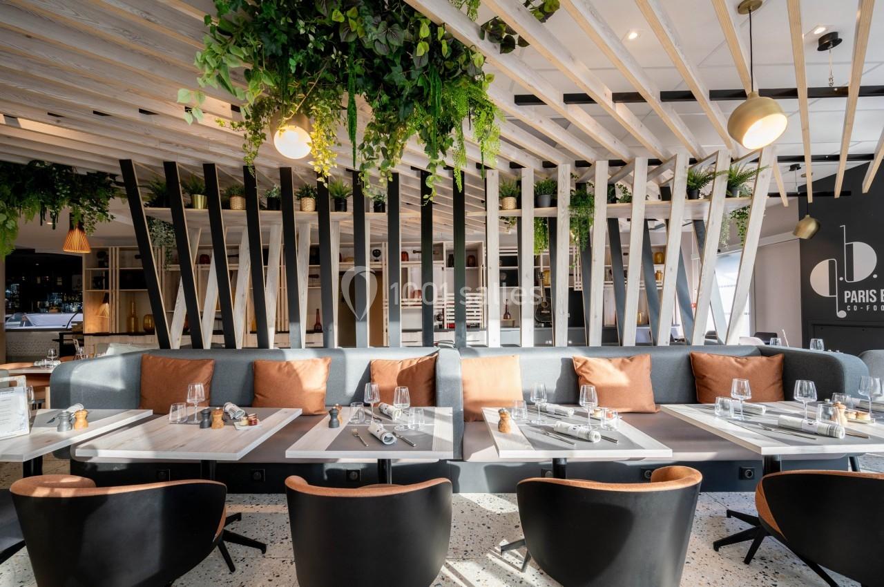 Salle de restaurant moderne avec tables dressées, banquettes grises, coussins orange et plantes suspendues.