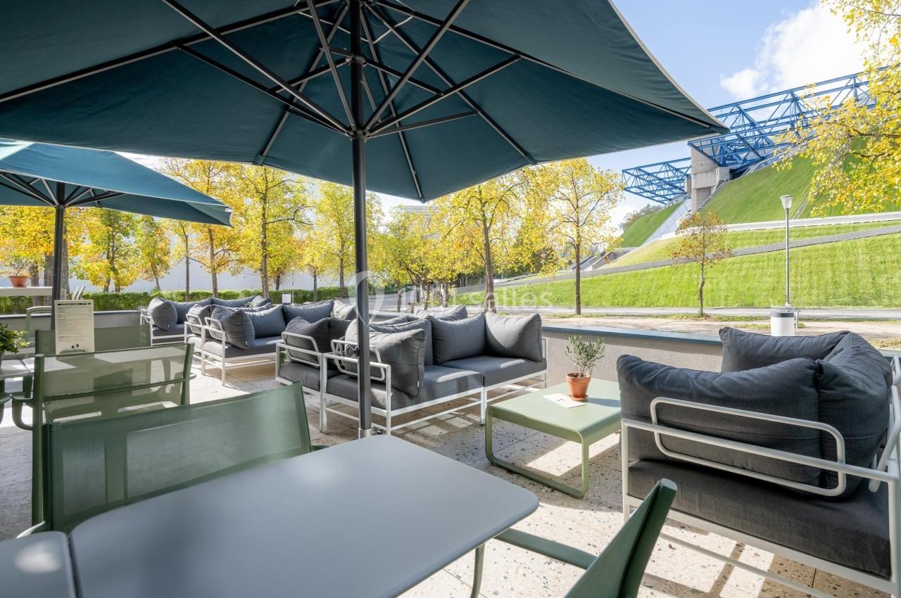 Terrasse extérieure avec tables, chaises et canapés sous des parasols, entourée d'arbres et d'une pelouse en pente.
