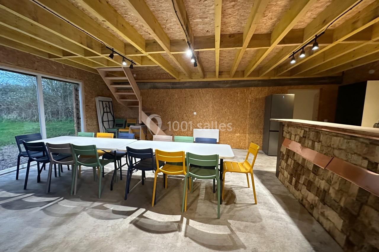 Salle avec une grande table entourée de chaises colorées, un escalier en bois et un bar en briques dans un espace lumineux.