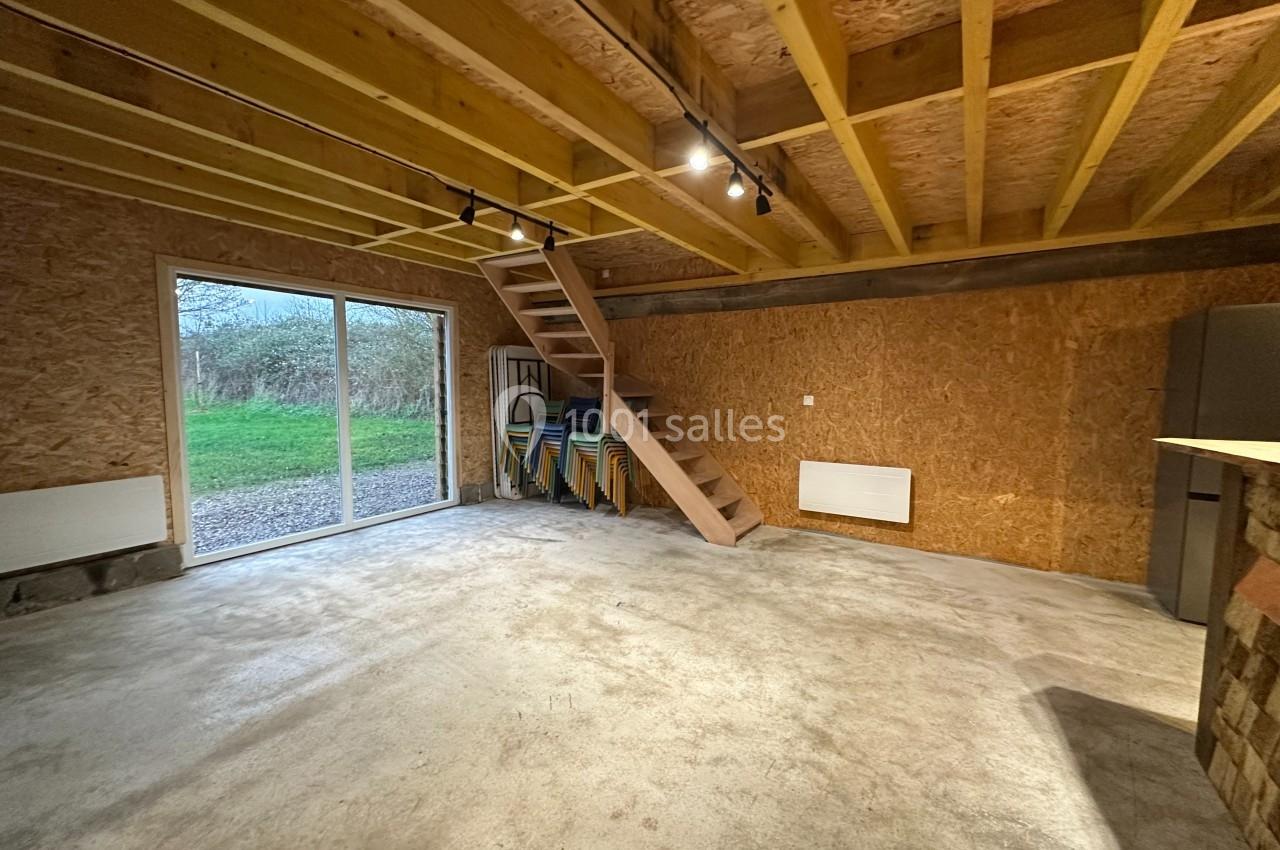 Intérieur d'une pièce avec sol en béton, murs en bois, escalier en bois, baie vitrée et chaises empilées dans un coin.