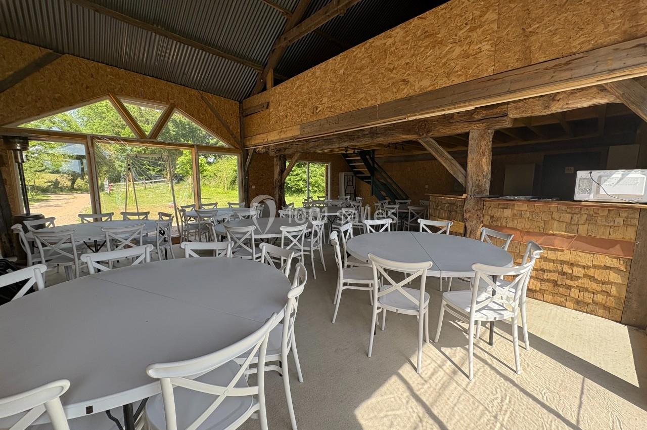 Salle lumineuse avec des tables rondes et des chaises blanches, charpente en bois apparente et grandes baies vitrées.