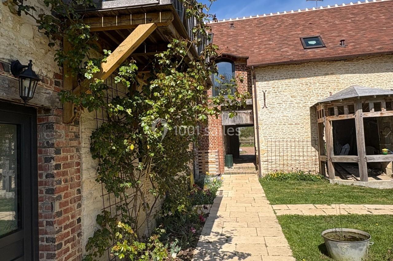 Cour pavée bordée de bâtiments en pierre avec balcon en bois, jardin verdoyant et ciel bleu en arrière-plan.