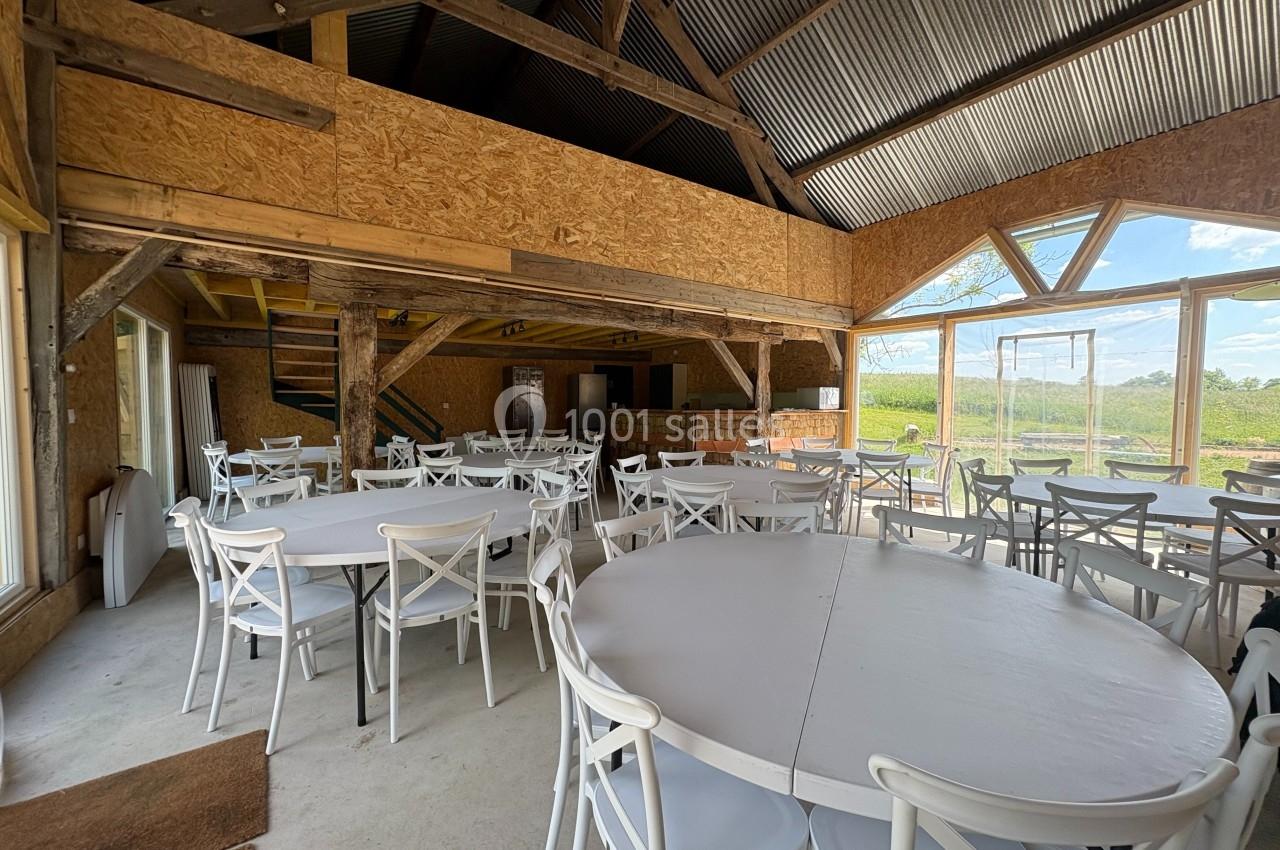 Salle lumineuse avec tables rondes et chaises blanches, poutres apparentes en bois et grandes fenêtres donnant sur l…