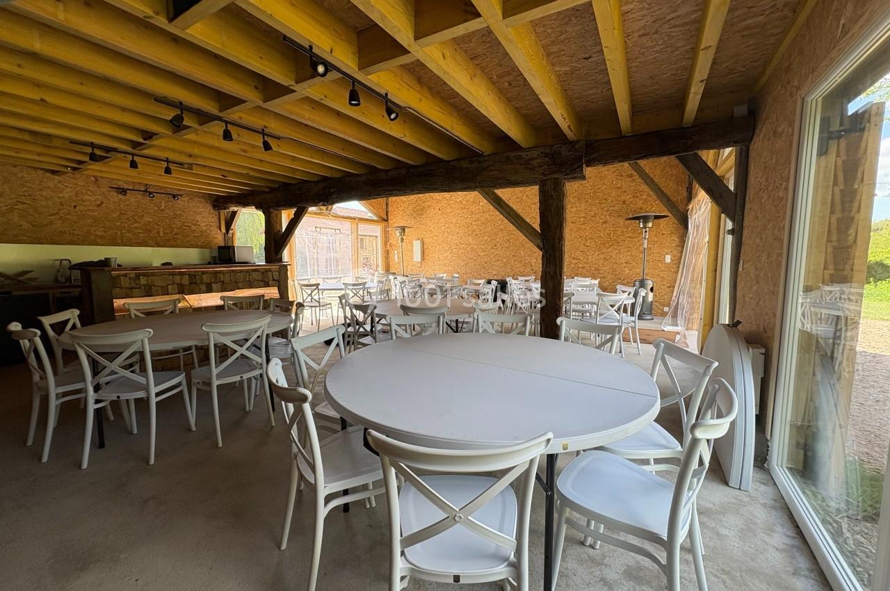 Salle avec des tables rondes et chaises blanches, charpente en bois apparente et vue sur une cour extérieure.