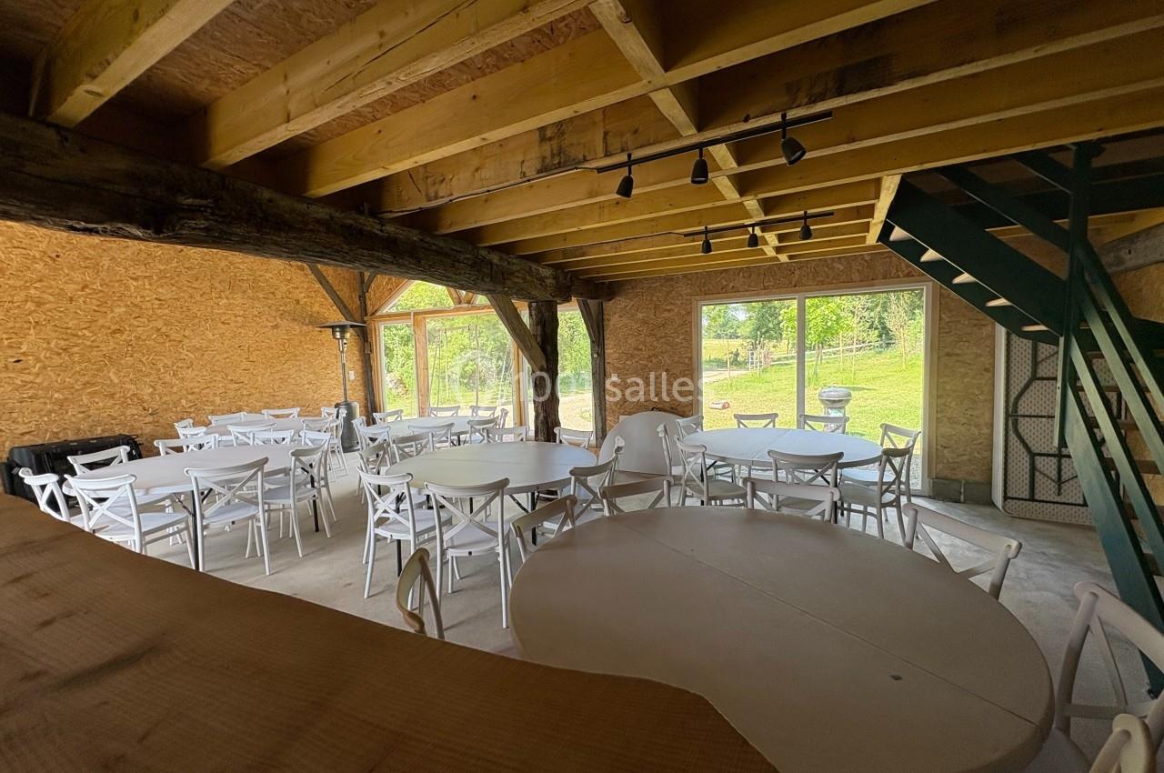 Salle en bois avec grandes baies vitrées, tables rondes et chaises blanches disposées pour un événement.