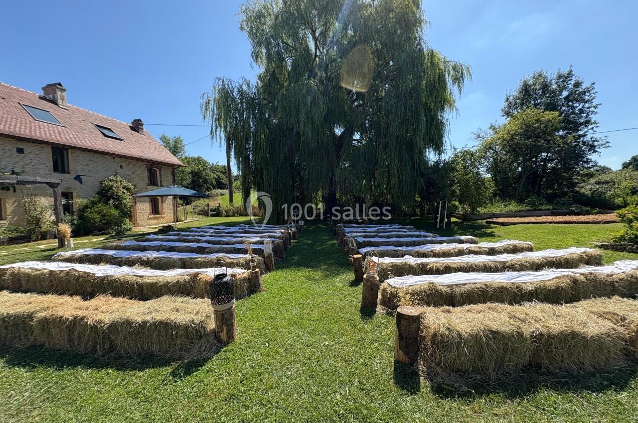 Rangées de bottes de foin recouvertes de tissu blanc disposées en extérieur sous un grand arbre, près d'une maison en pierre.