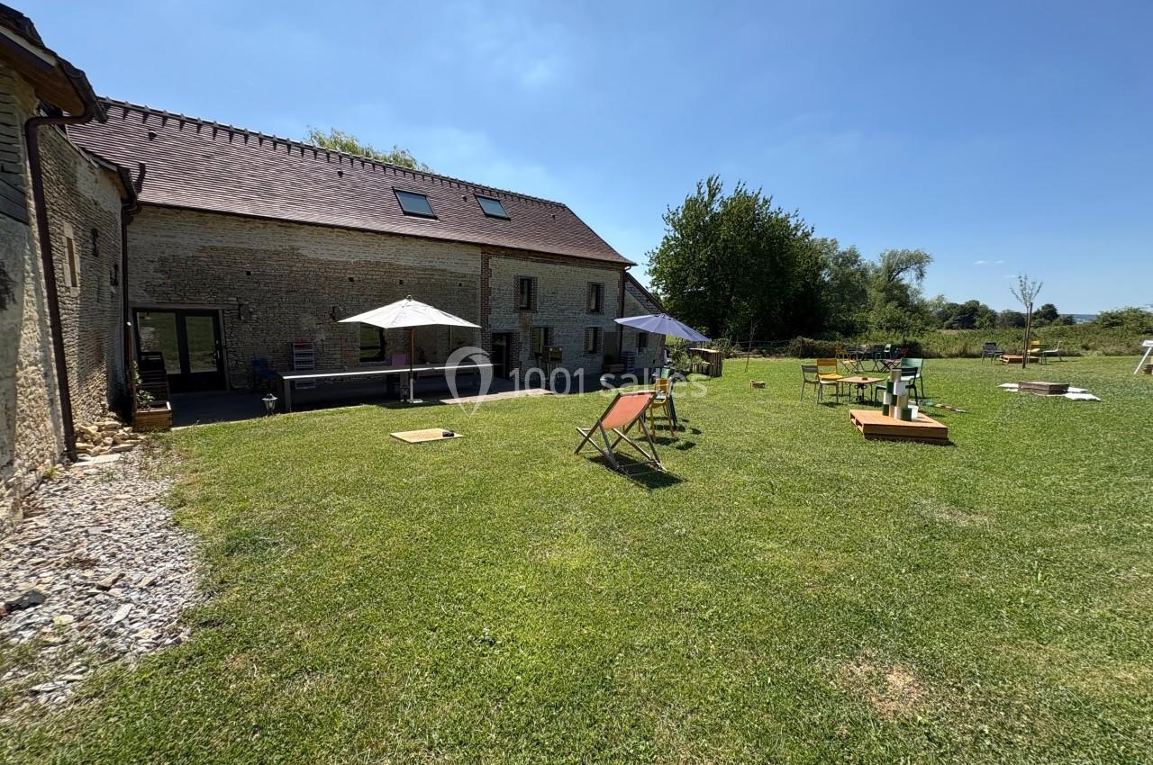 Jardin avec pelouse, chaises longues, parasols et jeux extérieurs devant une maison en pierre sous un ciel dégagé.