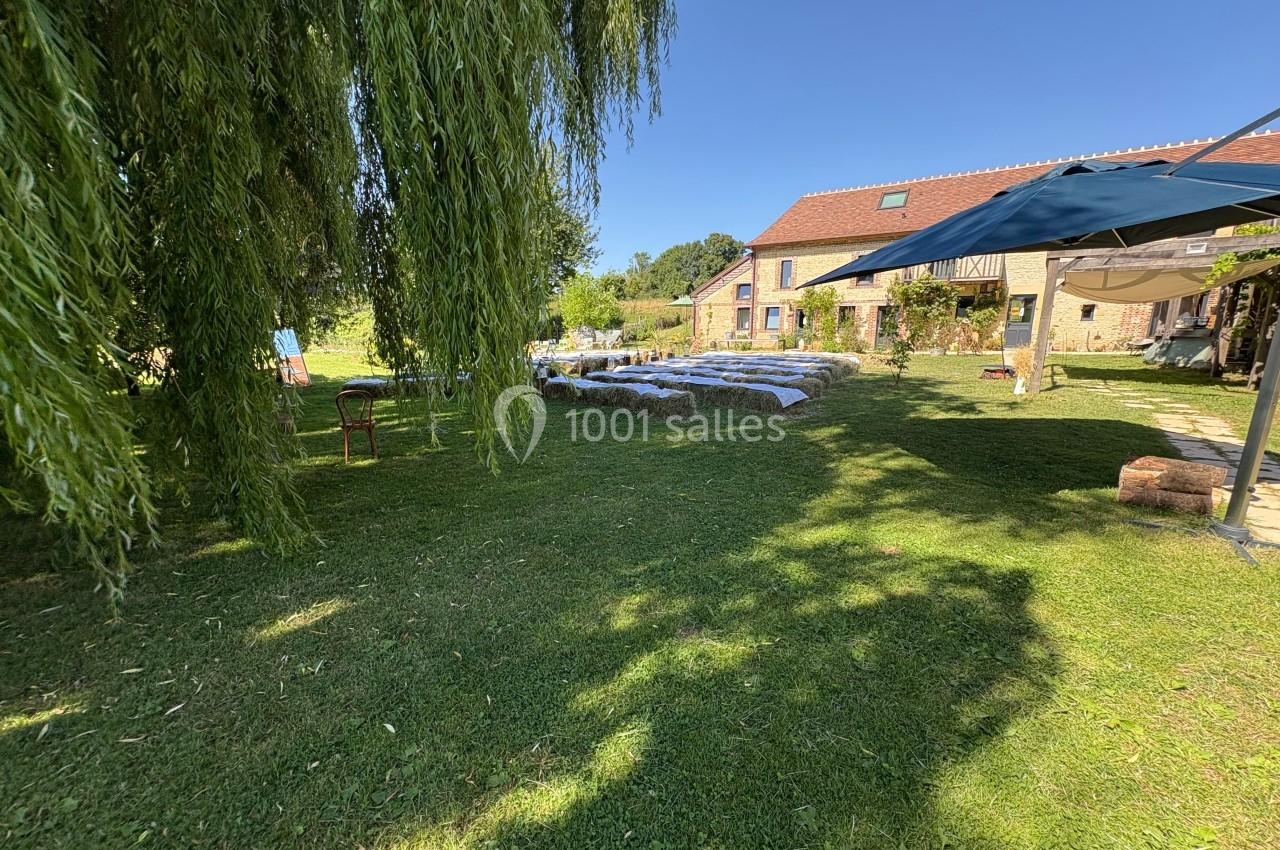 Jardin verdoyant avec un saule pleureur, des tables sous des parasols et une maison en pierre en arrière-plan.