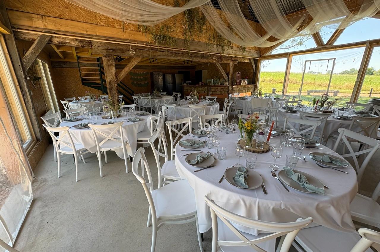 Salle de réception rustique avec tables rondes dressées, nappes blanches et décorations florales.