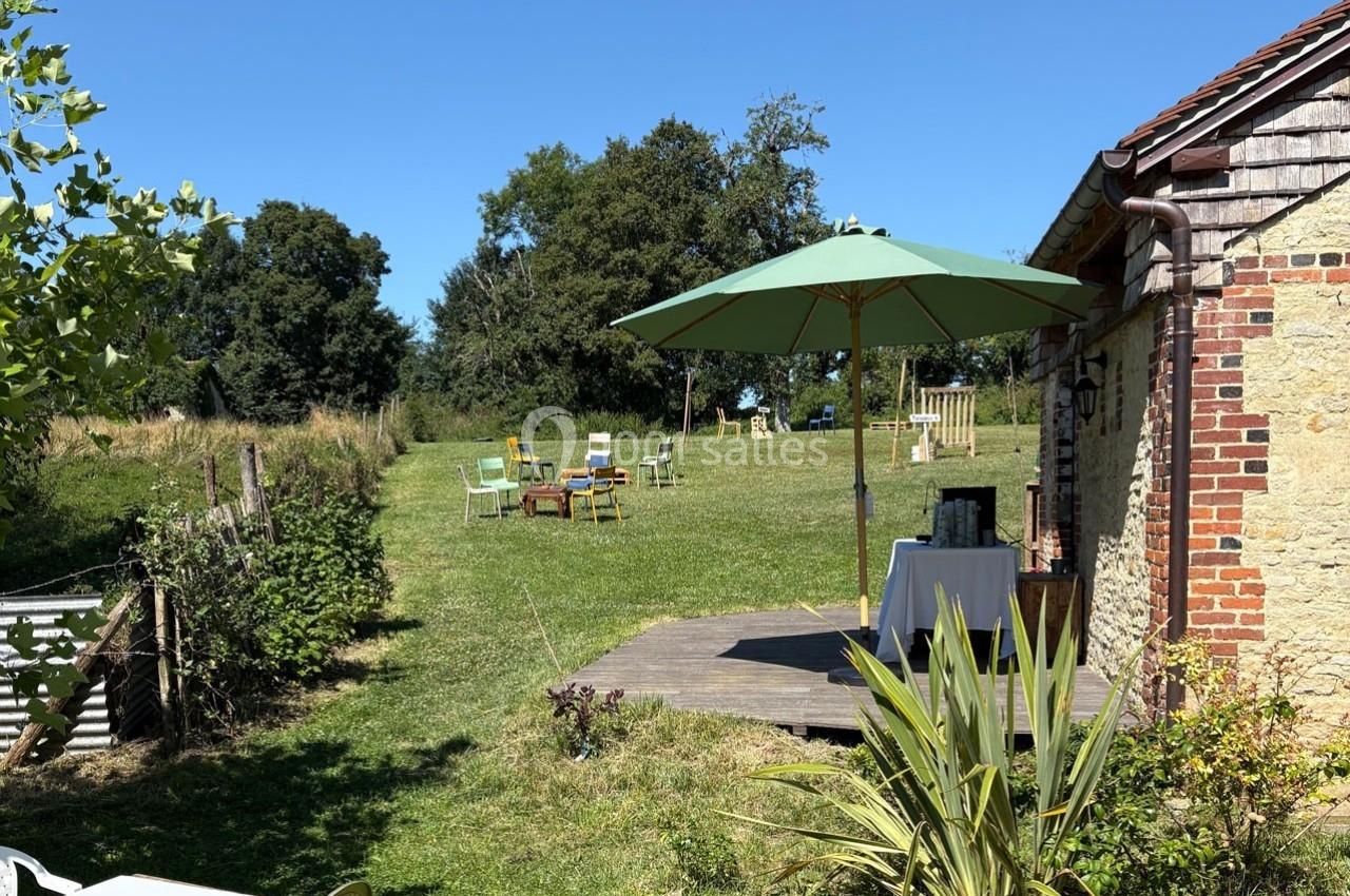 Jardin verdoyant avec des chaises colorées, une table sous un parasol vert et une maison en pierre à droite.