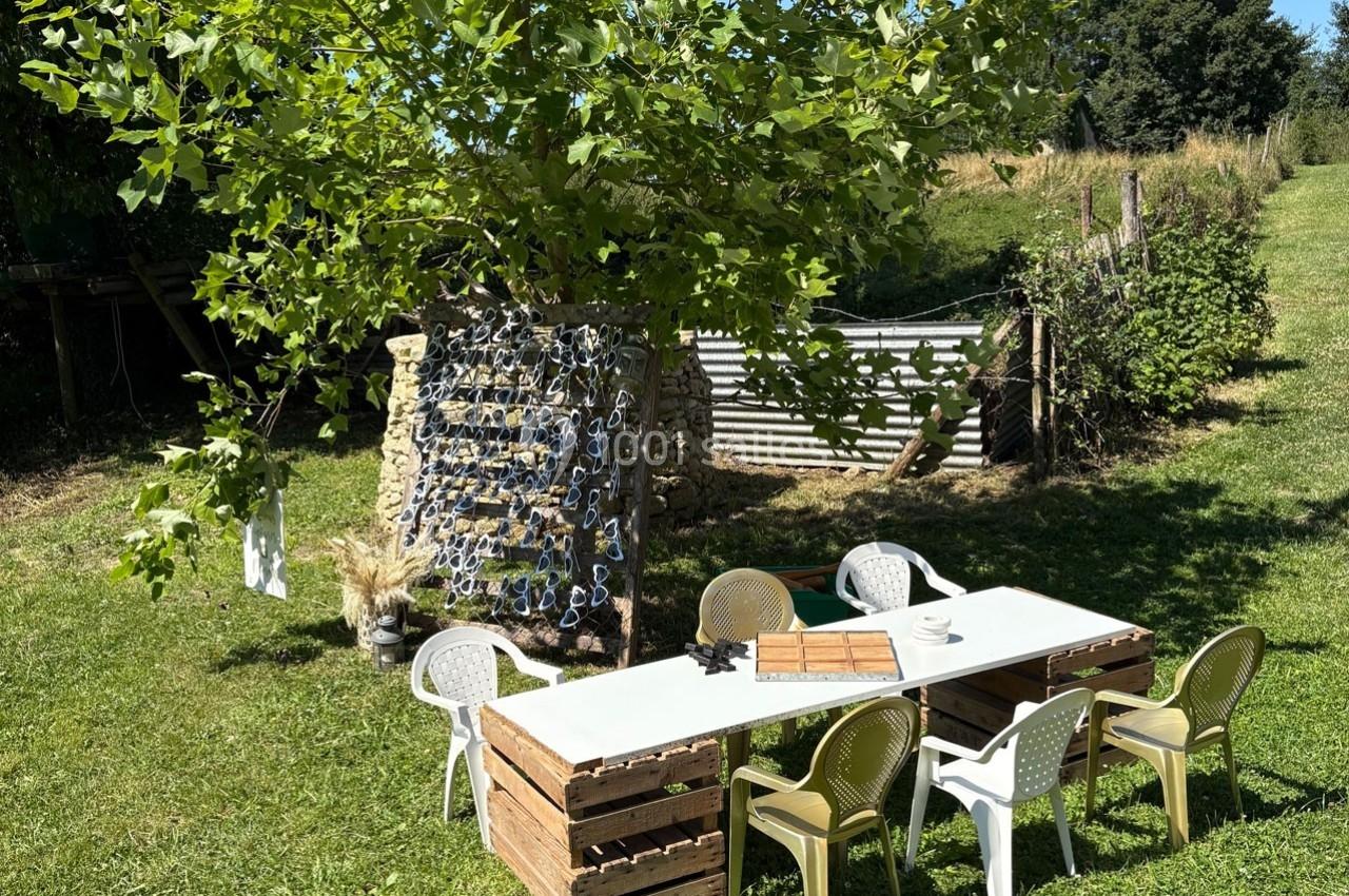 Table en extérieur sous un arbre, entourée de chaises, avec un décor champêtre et un panneau en arrière-plan.